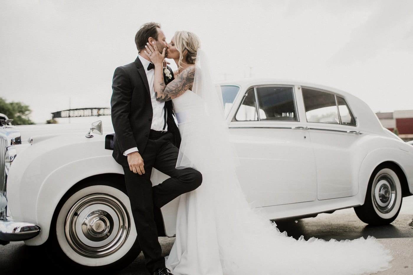 bride and groom kiss in front of white vintage car at Juniper Rooftop Wedding