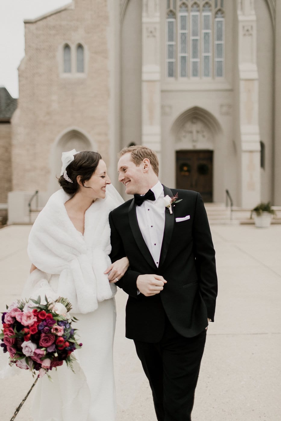 bridge and groom smile at each other in front of church at black tie winter wedding