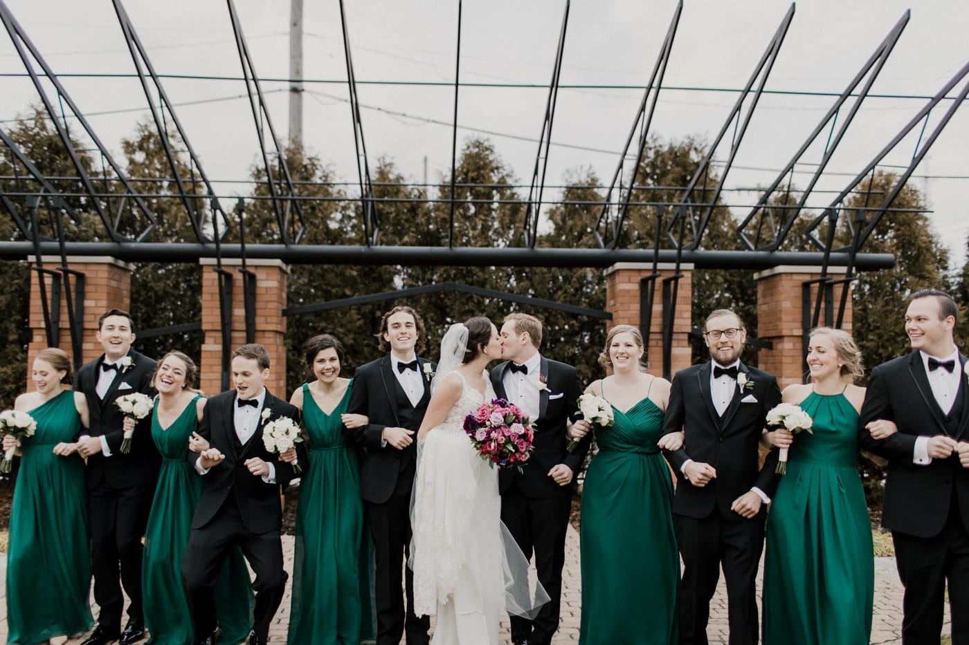 bride and groom kiss groomsmen and bridesmaids stand with arm interlocked at black tie winter wedding