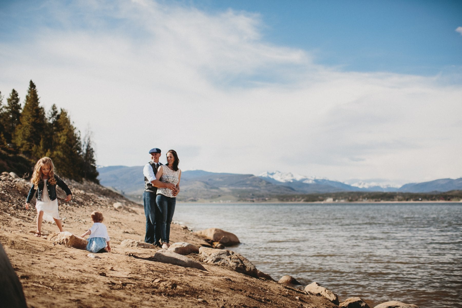 mom and dad embrace while kids play on lake shore at Grand Lake Colorado Family Portraits - Destination Photographer