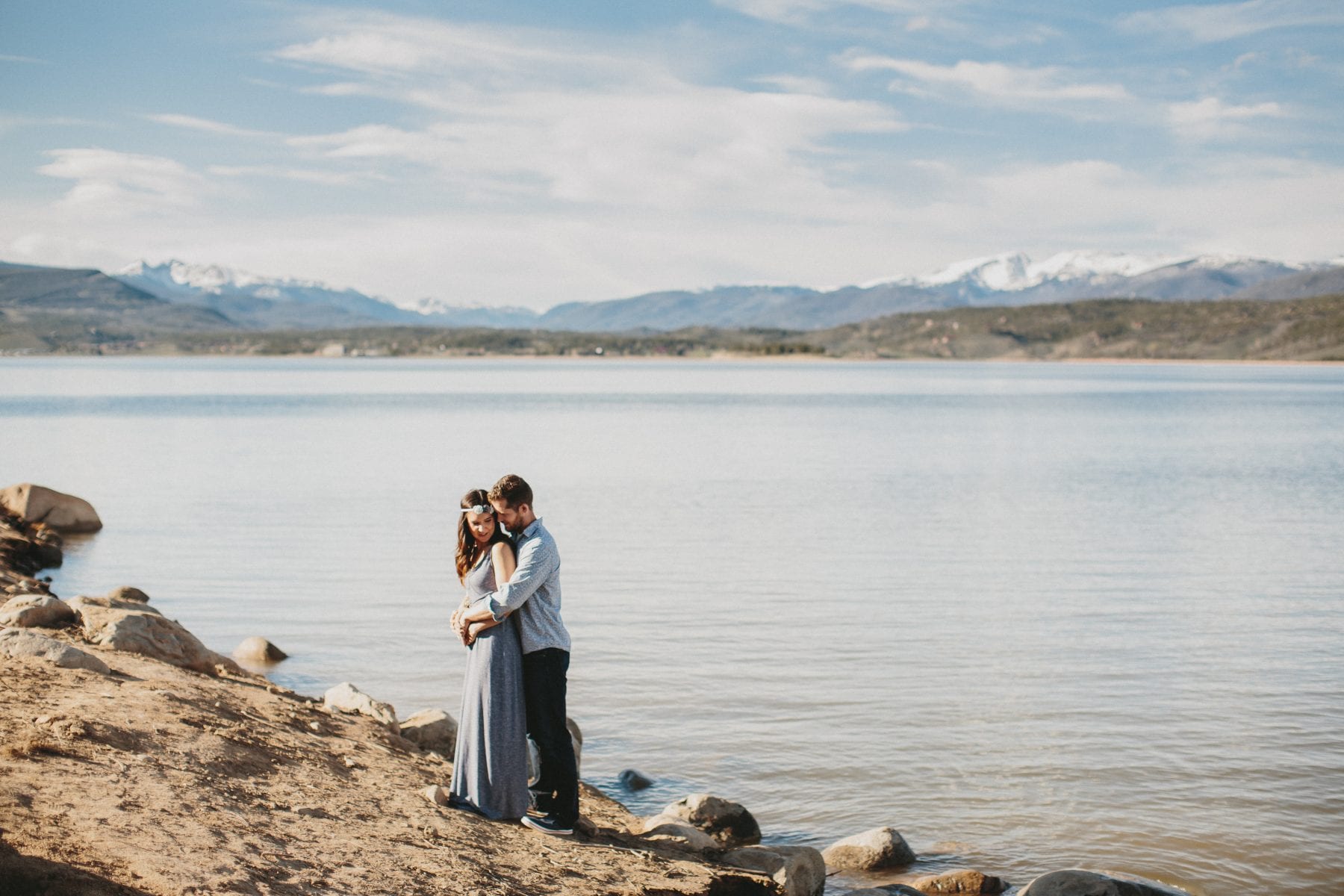 Couple embrace on lake shore with moutains in background - Grand Lake Colorado Family Portraits - Destination Photographer