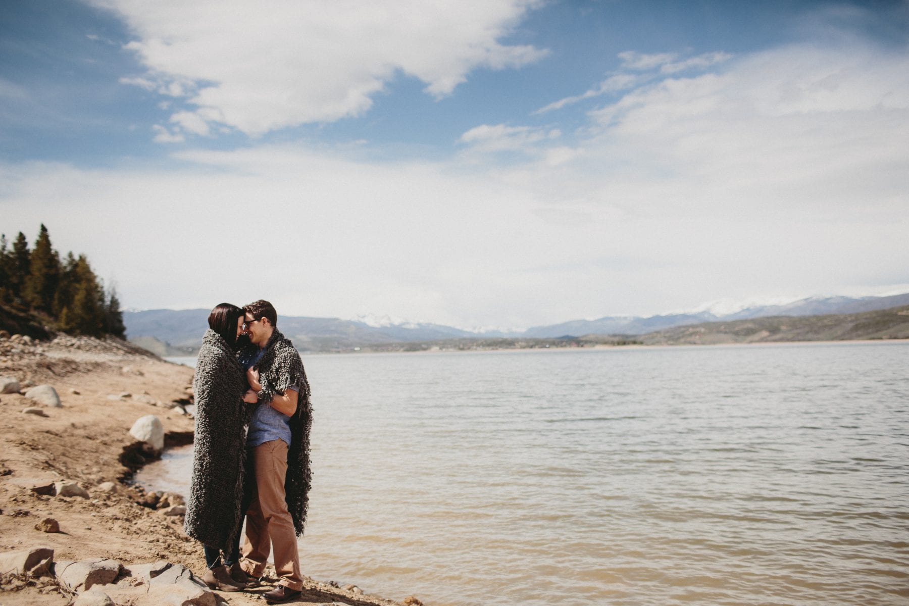 couple wrapped in blanket embraces on the lake shore at Grand Lake Colorado Family Portraits - Destination Photographer
